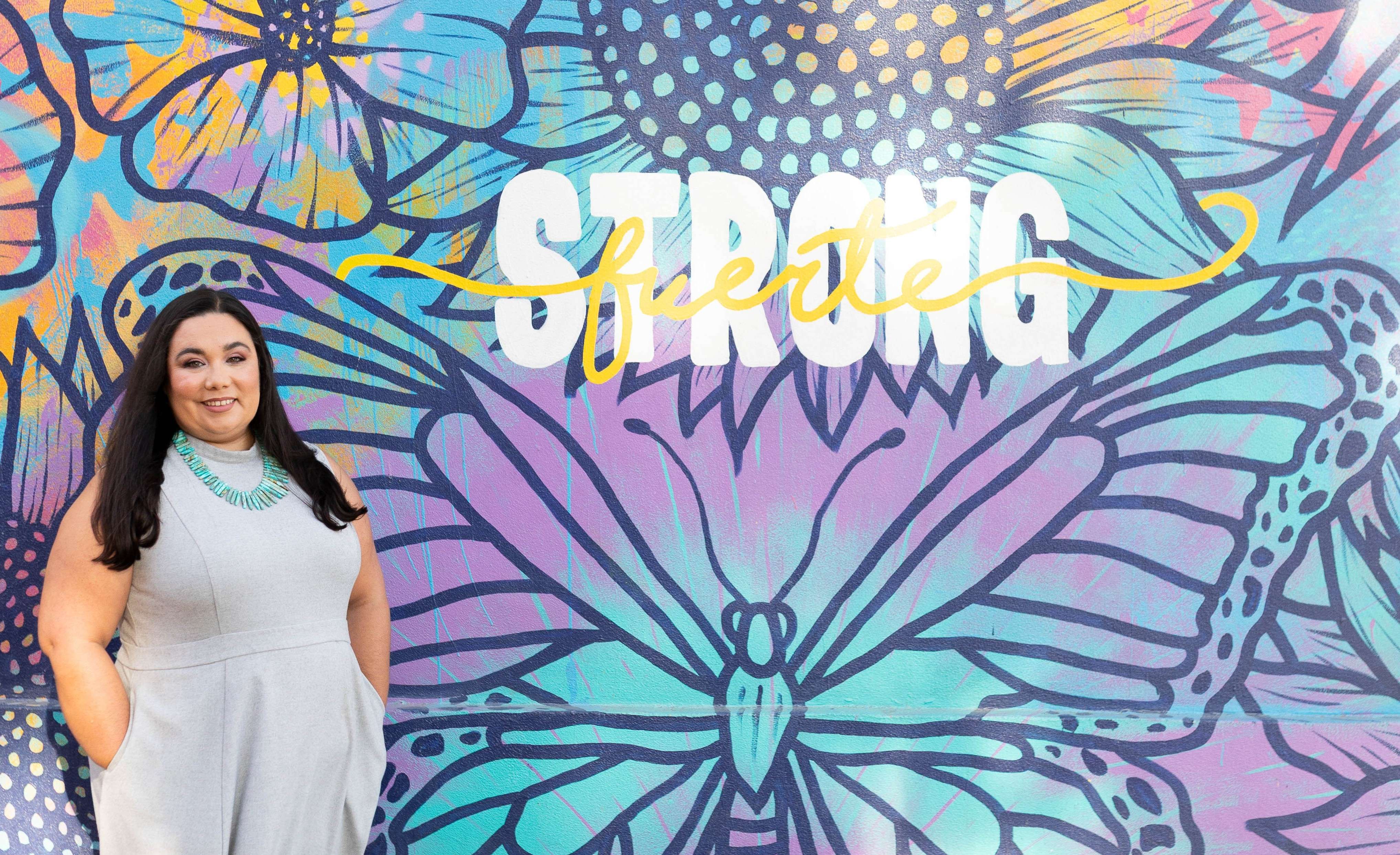 Elizabeth Ayala standing in front of a colorful butterfly mural with the words Strong and Fuerte